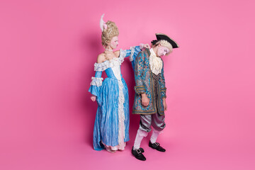 Elegant couple in vintage attire poses playfully against a pink backdrop. She in a baroque dress, he in a gentleman's costume