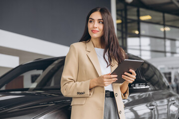 Smiling car seller, businesswoman in beige suit standing in car salon and using tablet for choosing right car.