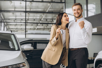 Happy young couple in casual clothes hugging and showing keys to new car in car showroom