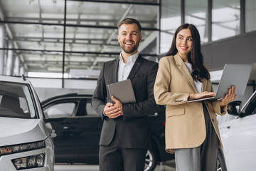 Super sales team in dealership, two consultants or managers in elegant suits with laptop and tablet in arms in car dealership