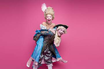 A joyful couple in elaborate baroque costumes poses against a vibrant pink background, embodying...