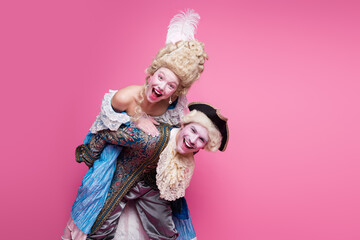 Elegant couple in baroque costumes against vibrant pink background, sharing joyful smiles and laughter