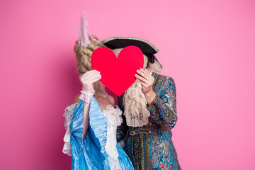 Couple in elegant baroque attire holding a heart, celebrating love against a pink backdrop during a...