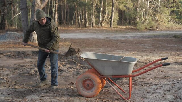 A person in a hooded jacket uses a shovel to dig the ground in a forested area while a wheelbarrow sits nearby on the dirt. The day appears clear and calm.