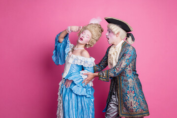 Elegant couple dressed in glamorous period costumes posing humorously against a pink background.