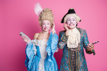 Charming couple in elegant baroque costumes pose playfully against a vibrant pink backdrop at a festive theme party.