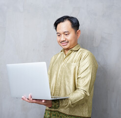 Adult Asian man wearing batik smiling at camera while using his laptop - Indonesia men