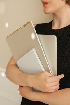 Close-up of a young woman in a smart black dress standing by a wall carrying a laptop computer, note pad and notebook