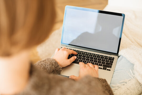 View from behind of a young woman sitting on a comfortable sofa in her living room using laptop computer with blank white screen