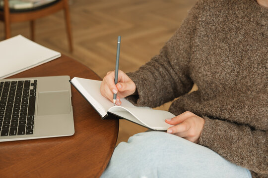 Close-up of a young woman sitting at her dining table using her laptop and writing in a note pad