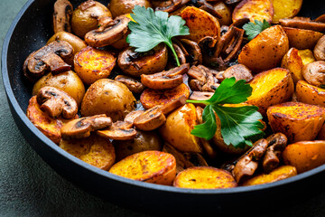 Boiled and fried baby potatoes in peels with fried champignon mushrooms and spices a black frying pan. Green background, top view
