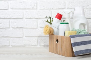 Eco-friendly cleaning products, supplies and green branches in crate on light wooden table against white brick wall, space for text