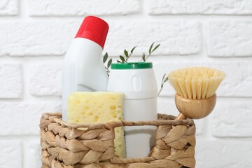 Eco-friendly cleaning products, supplies and green branches in wicker basket against white brick wall, closeup