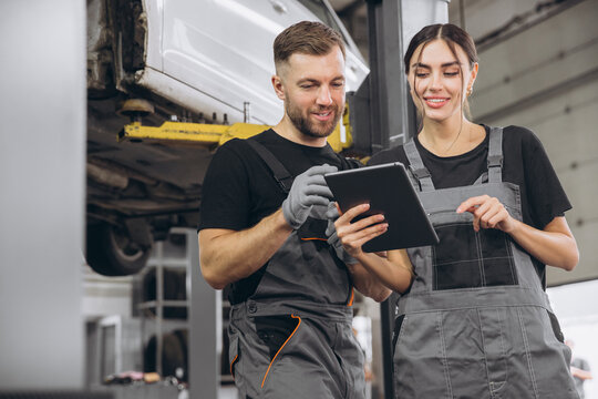 Young expert female inspects repair checklist with automotive mechanic worker partner, quality suspension technician team at fix garage. Vehicle maintenance service works industry occupation job.