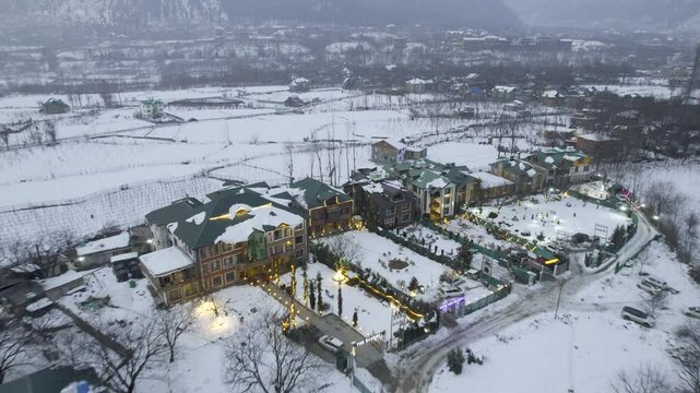 Aerial drone shot backward pan up reveal showing lit decorated houses in snow covered pahalgam rafting point valley with himalaya mountains and sunset sky in background in Kashmir India