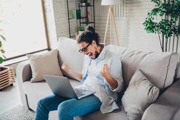 Excited young man celebrates online success while sitting on a comfortable sofa in a stylishly decorated living room during a bright day.