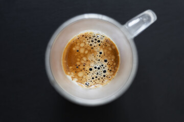 Top view of a freshly brewed espresso in a glass cup, showing frothy crema and bubbles on a dark background.