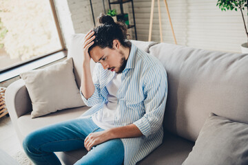 Young man indoors holding head while sitting on couch, thoughtful expression in casual setting,...
