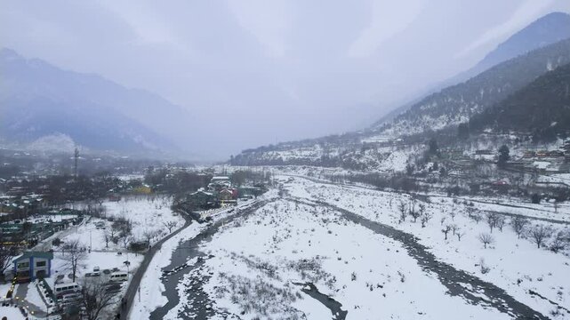 Aerial drone shot flying over lidder river with lit houses, hotels, resorts on the side towards himalaya mountains over the white snow covered landscape of Aru valley, Phelgam, Gulmarg, Sonmarg in