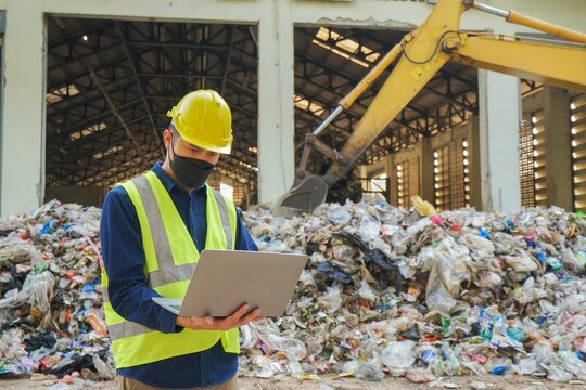 An environmental engineer uses a laptop to check the quality of a landfill in the background while a heavy machinery truck sorts waste at a waste plant.