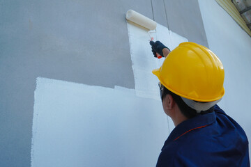 Asian male construction worker is painting with paint roller on white house wall, home decoration and construction concept in project.