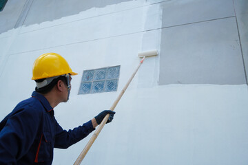 Asian male construction worker is painting with paint roller on white house wall, home decoration and construction concept in project.
