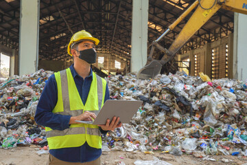 An environmental engineer uses a laptop to check the quality of a landfill in the background while a heavy machinery truck sorts waste at a waste plant.