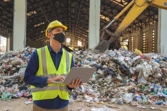 An environmental engineer uses a laptop to check the quality of a landfill in the background while a heavy machinery truck sorts waste at a waste plant.
