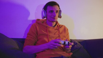 A man in a yellow hoodie in neon night light, puts on headphones, plays a game on a console with a joystick, and wins