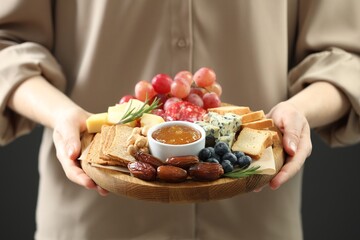 Woman holding board with different types of delicious cheese and other snacks on grey background, closeup
