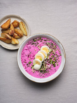 A vibrant bowl of Ukrainian beet soup features a creamy pink base topped with sliced eggs and herbs, alongside golden roasted potato wedges on a plate