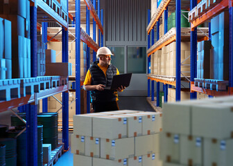 Man with laptop in factory warehouse. Enterprise storage area supervisor. Pallets with boxes on...
