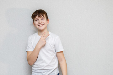 ten year old cute boy looking at camera standing against wall and making two finger gesture portrait