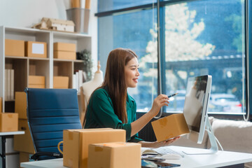 An Asian young woman sits in a room, working on a computer and making calls or video calls, managing her small business and online sales, surrounded by cardboard boxes ready for shipping.