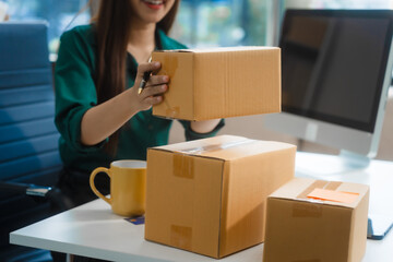 An Asian young woman sits in a room, working on a computer and making calls or video calls, managing her small business and online sales, surrounded by cardboard boxes ready for shipping.