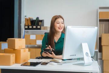 An Asian young woman sits in a room, working on a computer and making calls or video calls, managing her small business and online sales, surrounded by cardboard boxes ready for shipping.