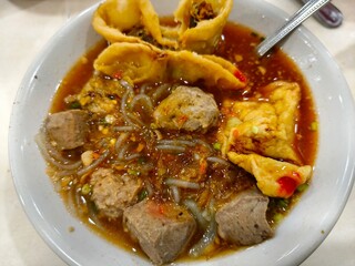 a bowl of bakso, a popular Indonesian meatball soup. The soup is filled with various types of meatballs, including beef and fish balls, as well as glass noodles and tofu puffs.