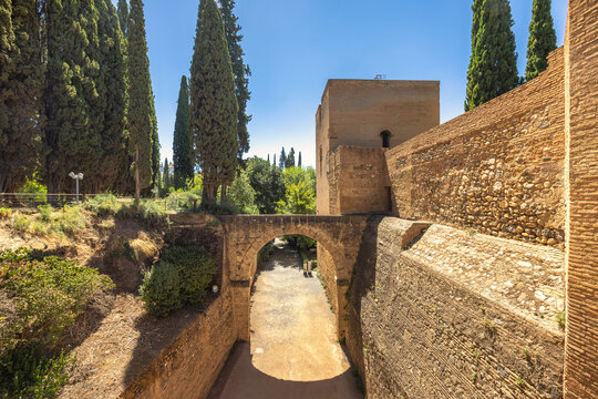 Aqueduct of the Acequia Real in the Alhambra complex, Granada town, Andalusia, Spain. Ancient stone archway and fortress walls under a clear blue sky. Tall cypress trees frame the scene.