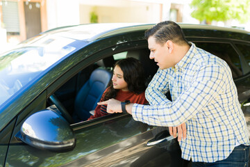 Latin dad teaching his teenage daughter how to drive, pointing at something inside the vehicle