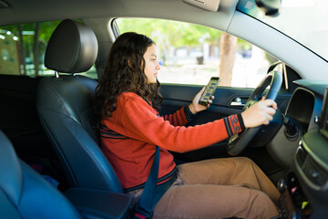 Hispanic teenager driving, glancing at her smartphone, highlighting dangers of distractions while driving and importance of safety measures like wearing a seatbelt