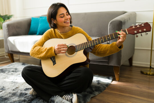 Happy hispanic woman sitting on the floor at home, playing her acoustic guitar and fully immersing herself in the joy of music