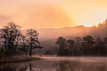 Dramatic atmospheric dawn landscape at Rydal Water in Lake District during Autumn with moody glow over mist on lake