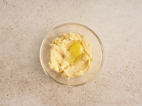 A glass bowl contains a smooth, creamy mixture with a fresh egg yolk placed at the center, indicating preparation for a baking recipe. The neutral background emphasizes the ingredients
