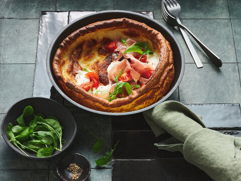 Freshly baked Yorkshire pudding layered with mozzarella, prosciutto, and vibrant cherry tomatoes sits on a sleek table. Fresh greens accompany the dish, enhancing the inviting presentation