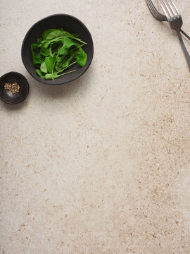 A black bowl filled with fresh green arugula sits beside a small dish of spices on a light-colored kitchen surface, ready for meal preparation