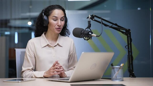 Businesswoman with a microphone and headphones is recording a podcast using a laptop sitting in a business office. Female coach conducts remote training. Vlogger communicates online via video call