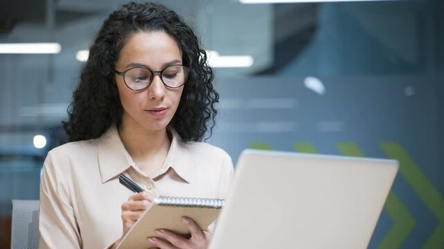 Businesswoman watching video call online conference taking notes looking at laptop screen sitting at workplace in office. Female employee listening to remote business training or course. Close up
