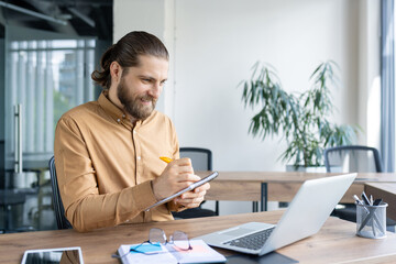 Smiling businessman working at a desk, writing in a notebook with a laptop nearby. Ideal for concepts of productivity, business planning, and professional work environments.