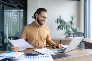 Fototapeta premium Successful individual concentrated on reviewing paperwork while using a laptop in a bright office. He appears professional, engaged, and content as he manages work efficiently.