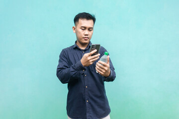 Asian man wearing navy shirt using smartphone and holding drink bottle isolated on green background